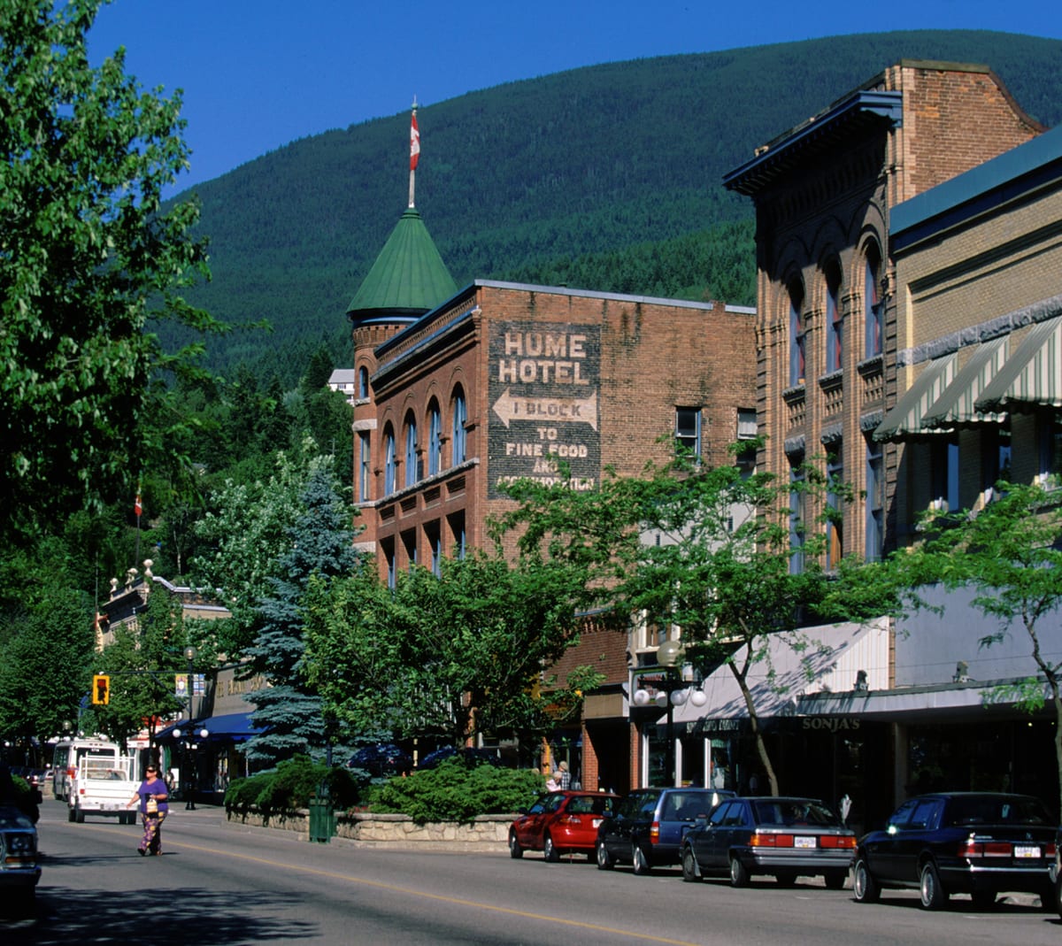 Baker Street in Nelson: brick heritage buildings with a green turret and a painted hotel sign, parked cars below and a forested mountain behind.