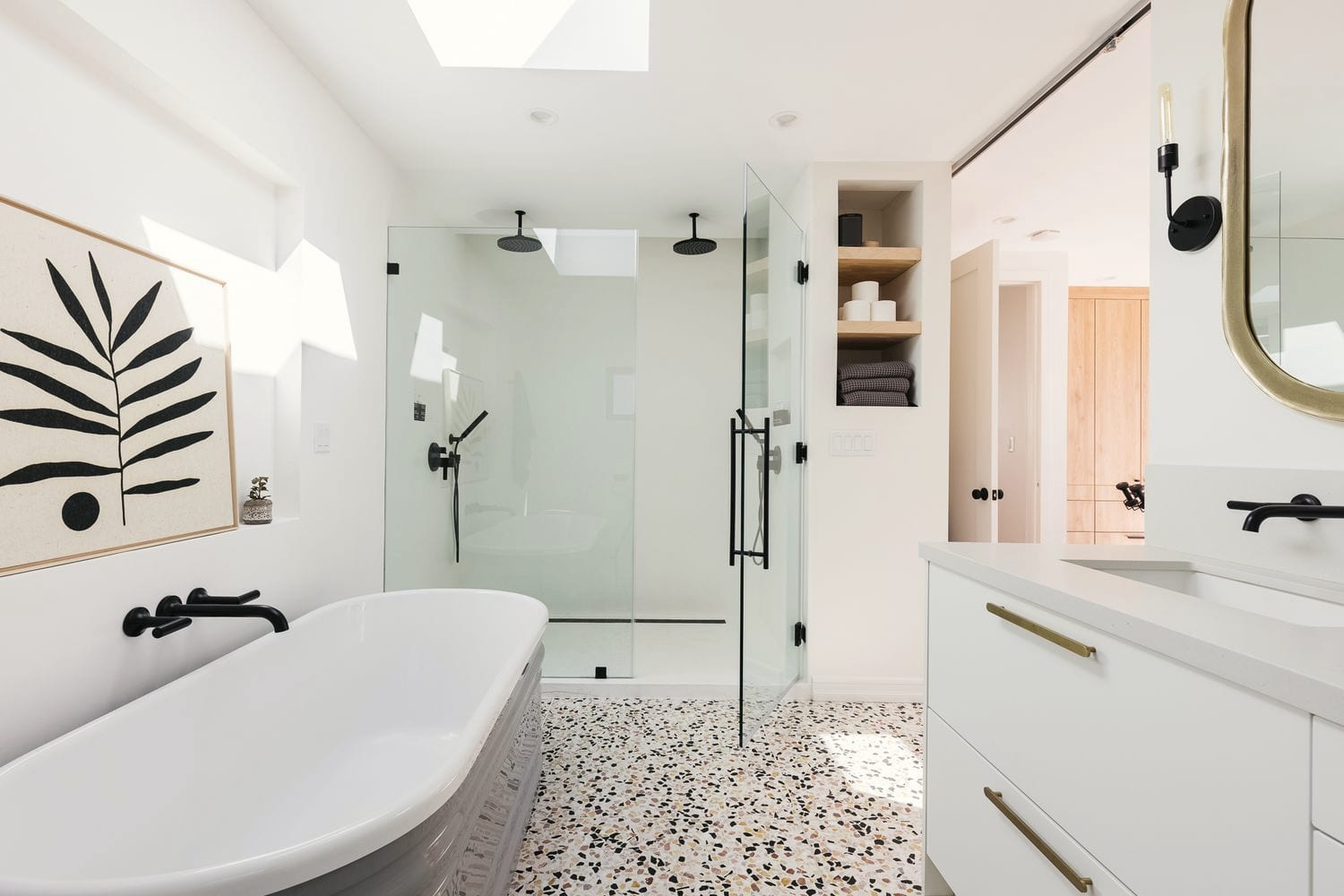 A bright bathroom with a frameless glass shower, a freestanding tub and a terrazzo floor under a skylight.