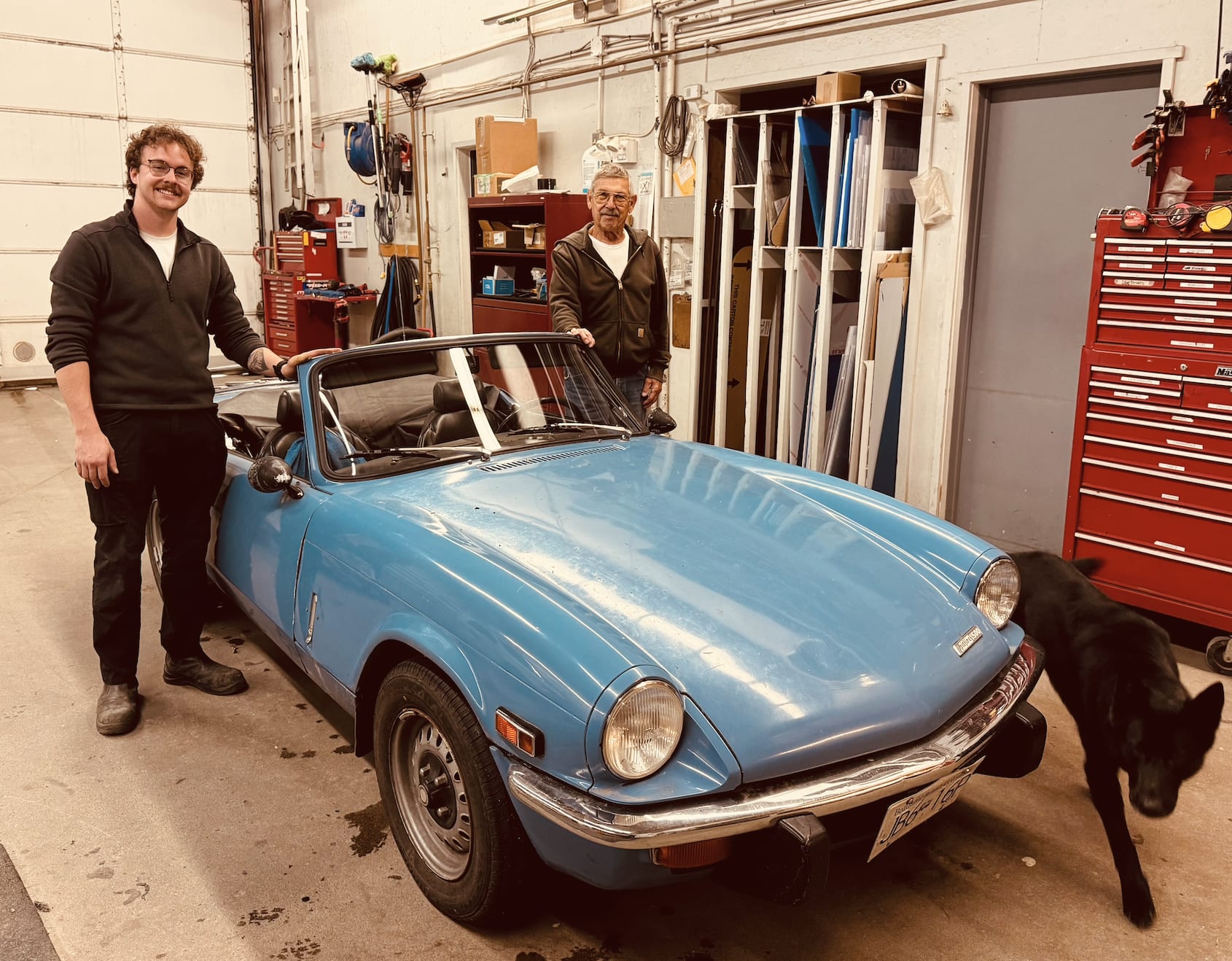 A light-blue classic convertible in the shop bay, a man standing at each end of it, and a black dog walking past the front bumper.