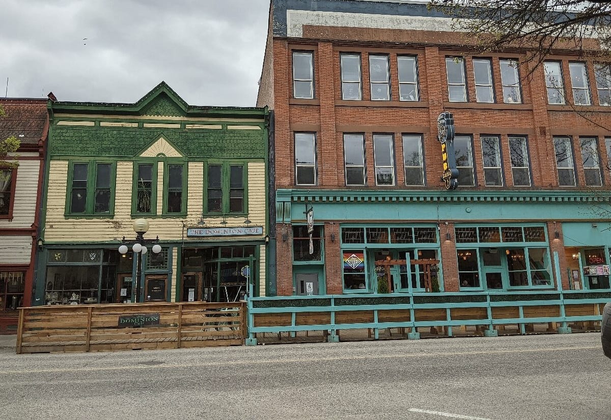 Heritage storefronts seen from across the street: a green and cream wooden front beside a red brick block, both glazed in tall shop windows above patio railings.