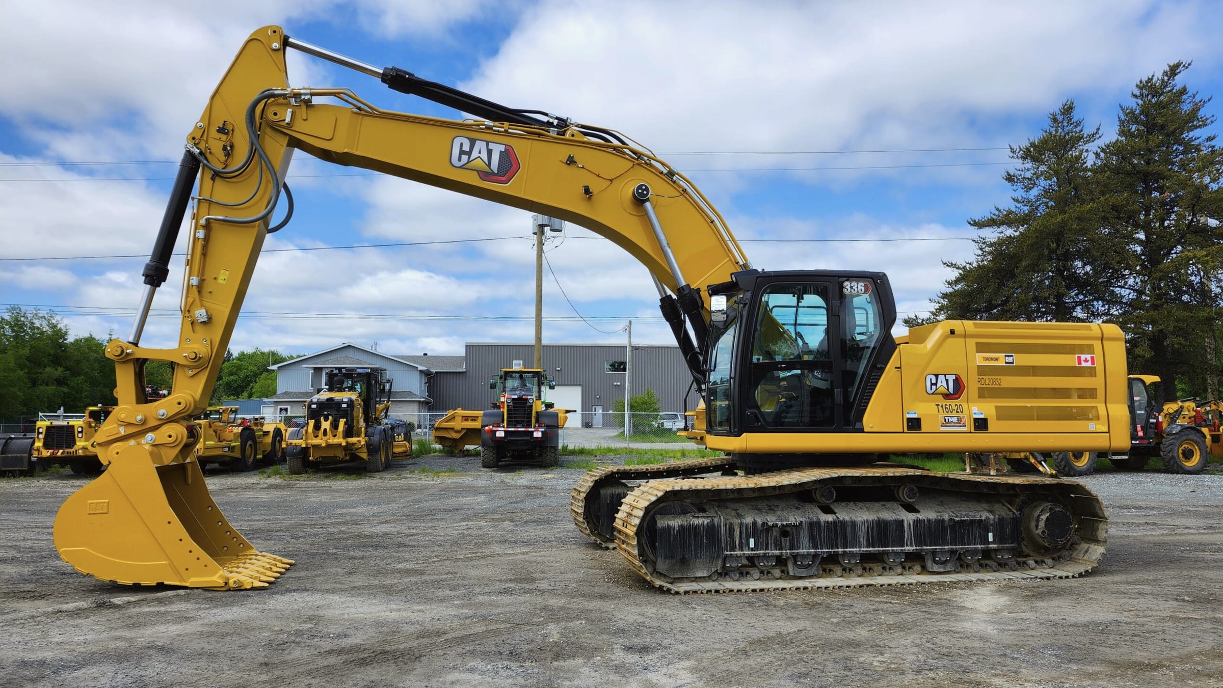 A yellow tracked excavator with its bucket lowered, parked in a gravel yard with other yellow machines behind it.