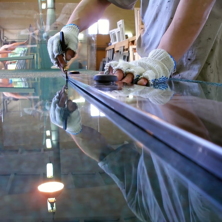 Gloved hands scoring a sheet of glass with a cutter along a straightedge, the workshop reflected in the glass.