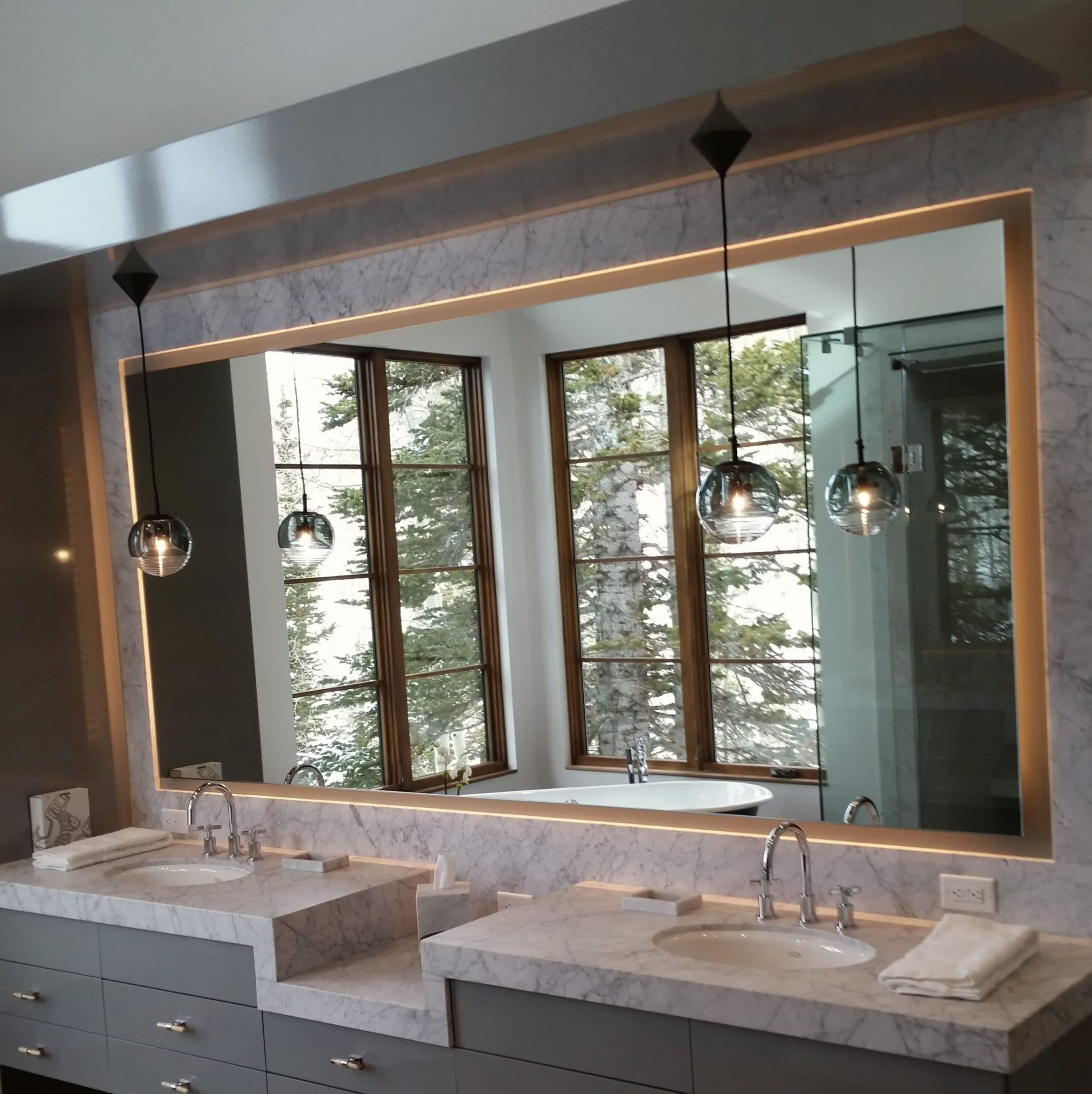 A wide lit mirror over a double marble vanity, reflecting two windows and pendant lights.