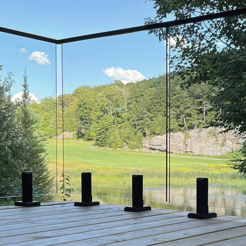 A frameless glass railing on black posts along a wooden deck, with a green field and trees beyond.