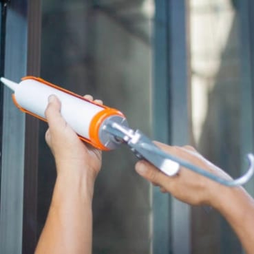 Two hands holding a caulking gun, a tube of sealant loaded, against a glass and metal frame.