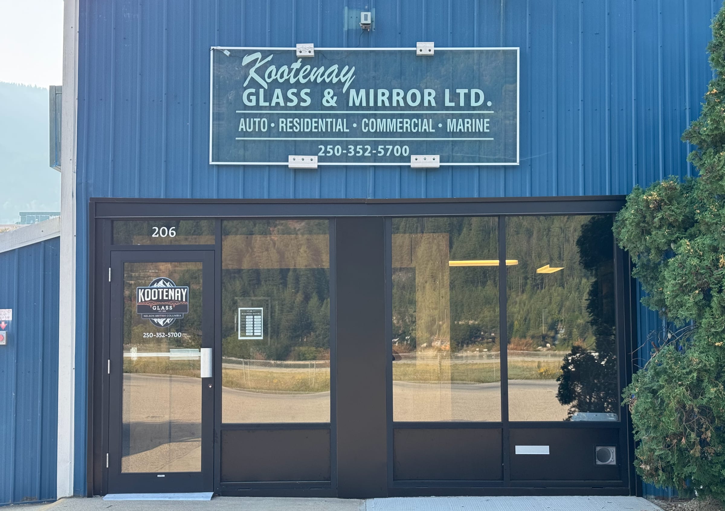 The blue steel front of the shop at 206: a glass sign lettered Kootenay Glass & Mirror Ltd. for auto, residential, commercial and marine glass, over a dark-framed glass storefront and entrance door.