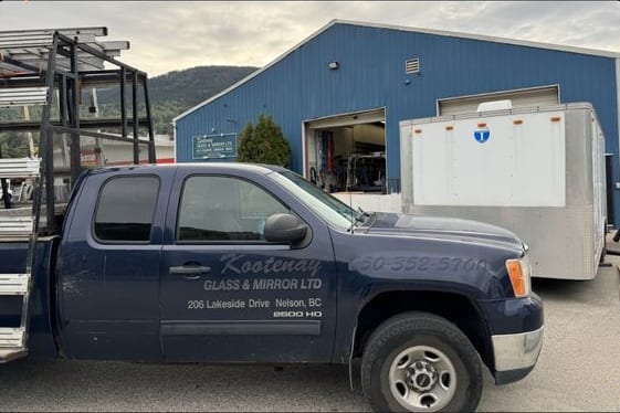 A Kootenay Glass & Mirror pickup with a glass rack, lettered with the shop's phone number and 206 Lakeside Drive, parked outside the blue shop with the bay door open.