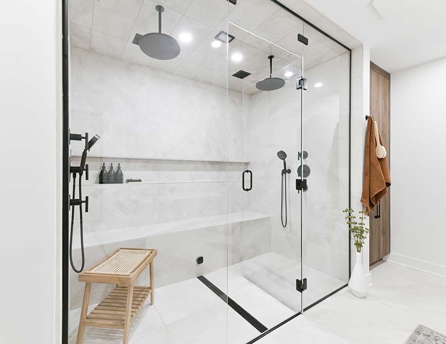 A large walk-in shower behind frameless glass with black hardware, a teak bench inside and two rain heads overhead.