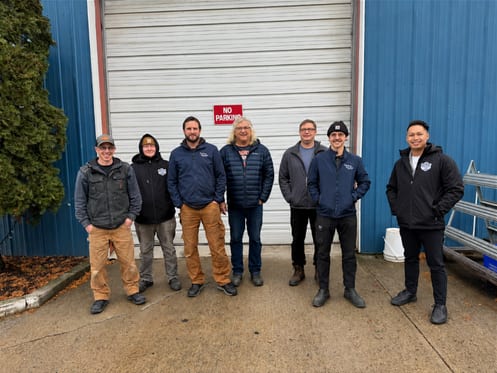 The Kootenay Glass team, seven people standing in a row in front of a white bay door.