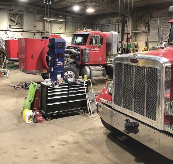 Two red heavy trucks in a repair shop, one with its hood up, beside a tool chest, with the grille of a third truck in the foreground.