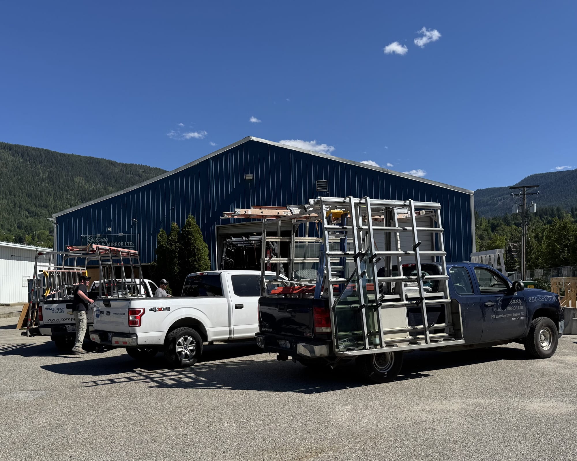 Three work trucks with glass racks parked outside the blue shop building, with forested mountains behind.
