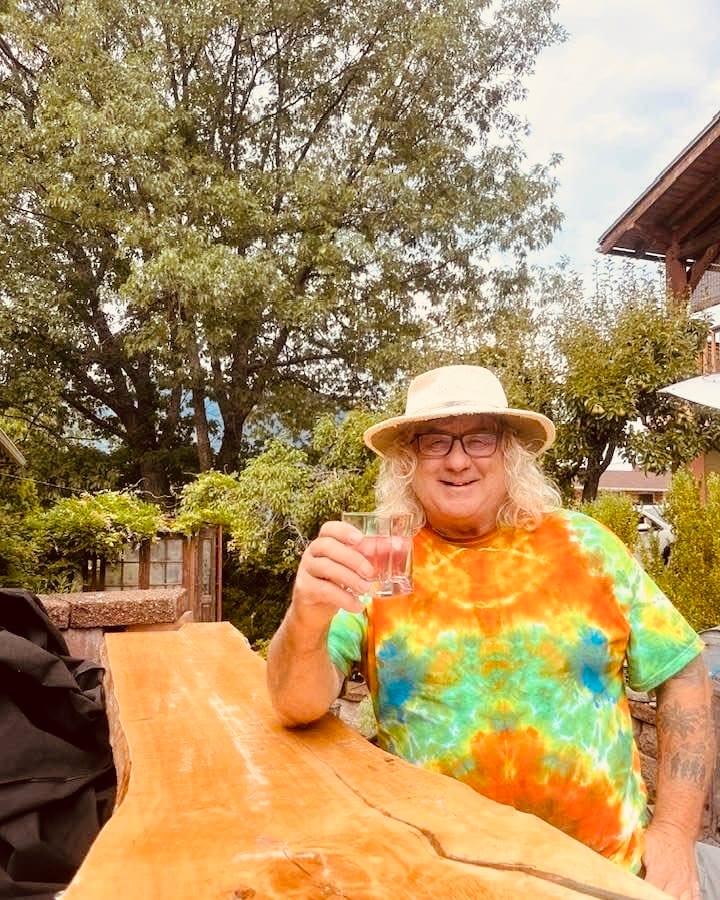A man with long white hair, a straw hat and glasses, in a tie-dye shirt, raising a glass at a live-edge wood table outdoors under tall trees.